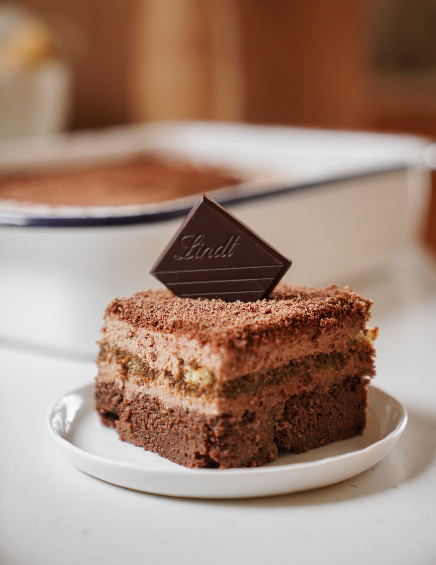 A piece of layered chocolate cake topped with a square of Lindt dark chocolate sits on a small white plate, with a baking dish of more cake blurred in the background.
