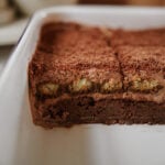 A close-up of a rectangular chocolate layered dessert in a white baking dish, showing distinct layers of chocolate, cream, and sponge cake with a dusting of cocoa powder on top. A portion has been cut out.