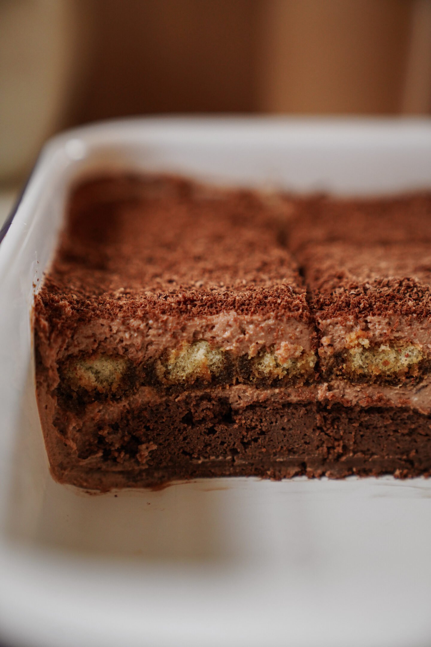 A close-up of a chocolate dessert in a white dish, featuring visible layers of chocolate cake, cream filling, and cocoa powder on top, with a portion already served, revealing the inside texture.