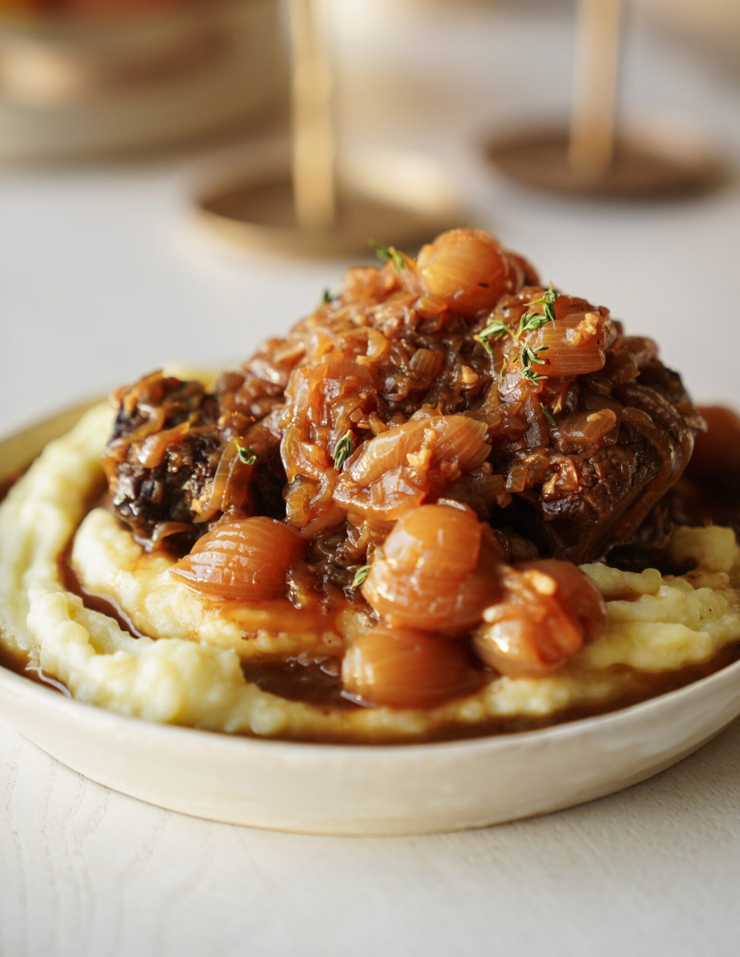 A close-up of braised meat topped with caramelized onions and pearl onions, served on a bed of creamy mashed potatoes in a shallow bowl.