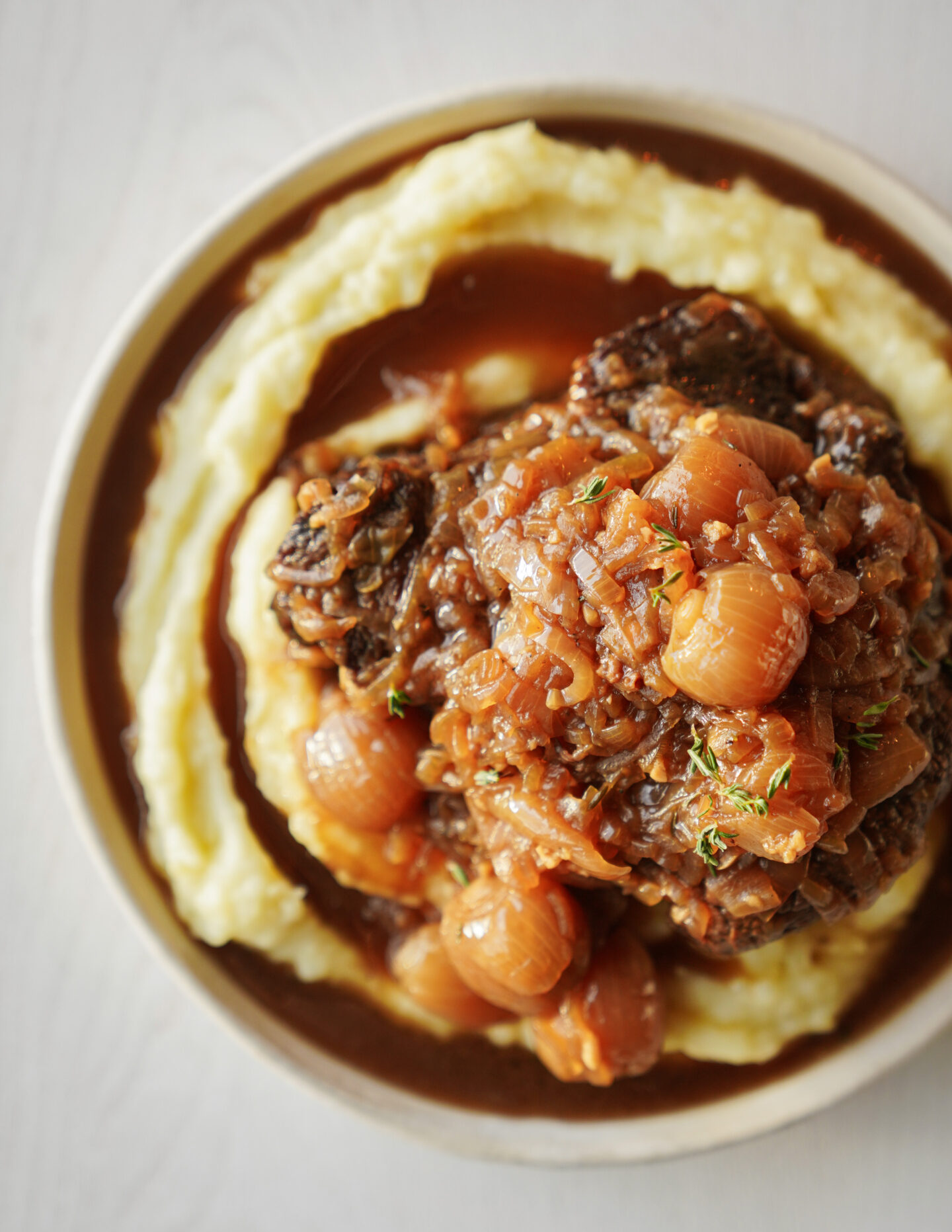 A plate of mashed potatoes topped with braised beef and pearl onions in a rich brown sauce, garnished with fresh herbs. The dish is photographed from above on a neutral background.