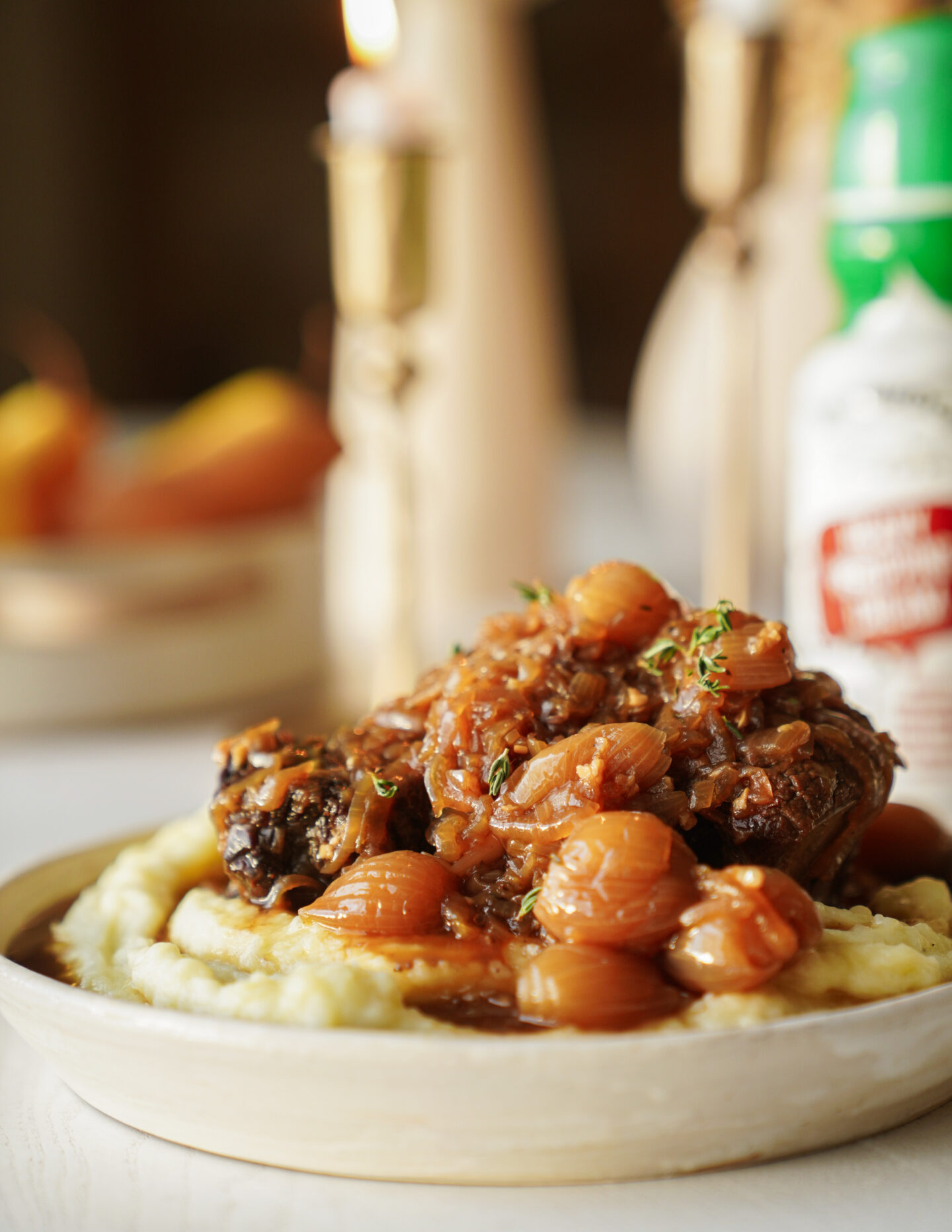 A close-up of a dish featuring braised meat topped with pearl onions and sauce, served over creamy mashed potatoes on a beige plate, with blurred candles and a bottle in the background.