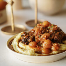 A bowl of creamy mashed potatoes topped with braised meat, pearl onions, and a rich brown sauce, garnished with fresh herbs, on a light-colored table.