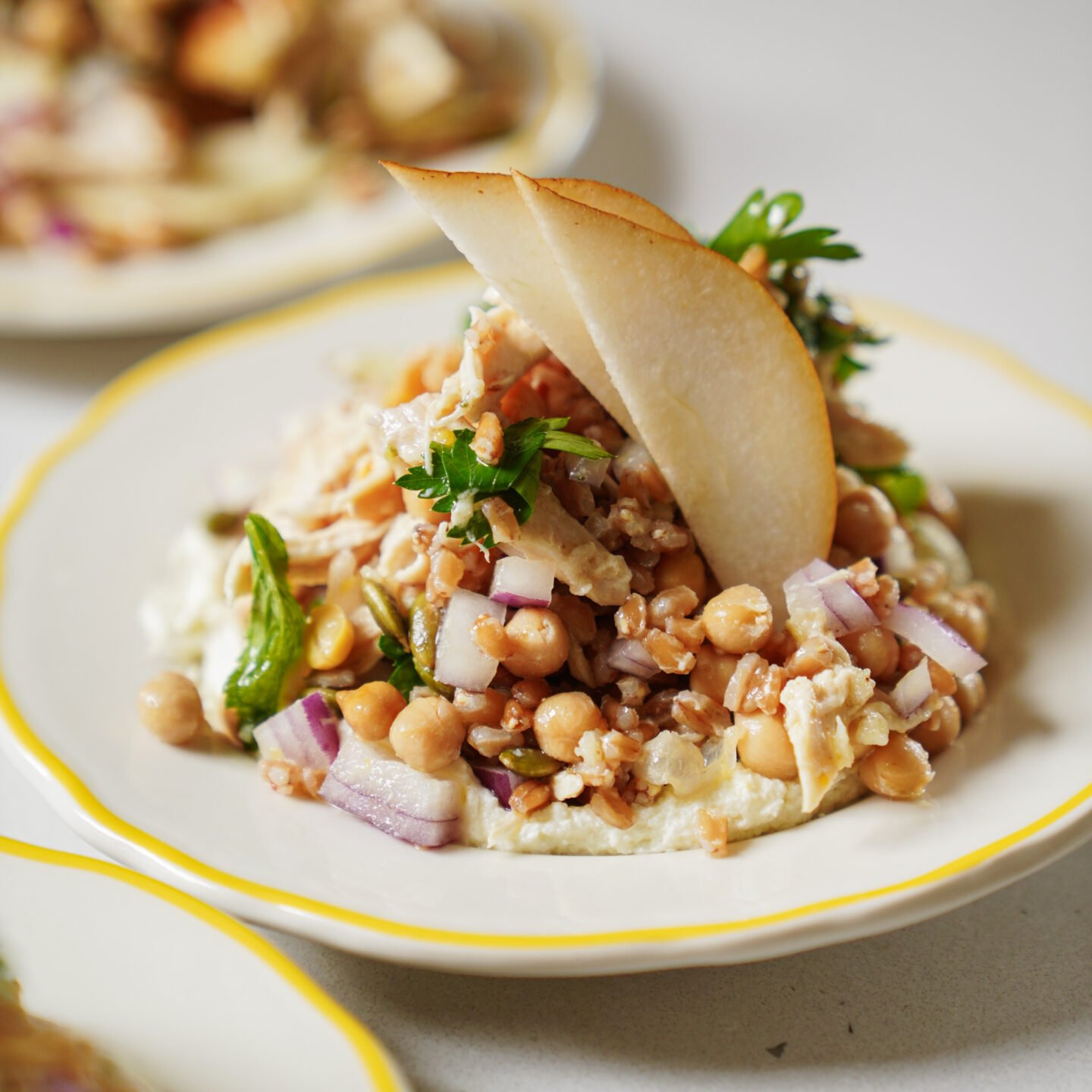A salad featuring chickpeas, red onion, chopped herbs, grains, and shredded chicken sits on a plate, garnished with two thin pear slices and fresh parsley.