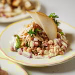 A salad featuring chickpeas, red onion, chopped herbs, grains, and shredded chicken sits on a plate, garnished with two thin pear slices and fresh parsley.