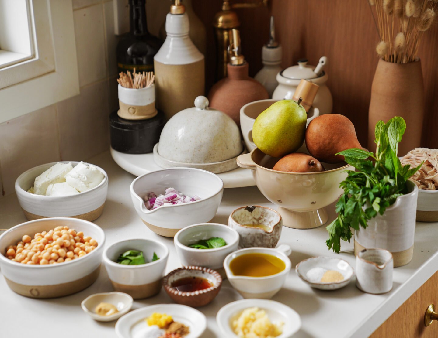 A kitchen counter displays bowls of chickpeas, sliced onions, spinach, olive oil, spices, cheese, and whole pears, with fresh herbs and various ceramic containers in the background.