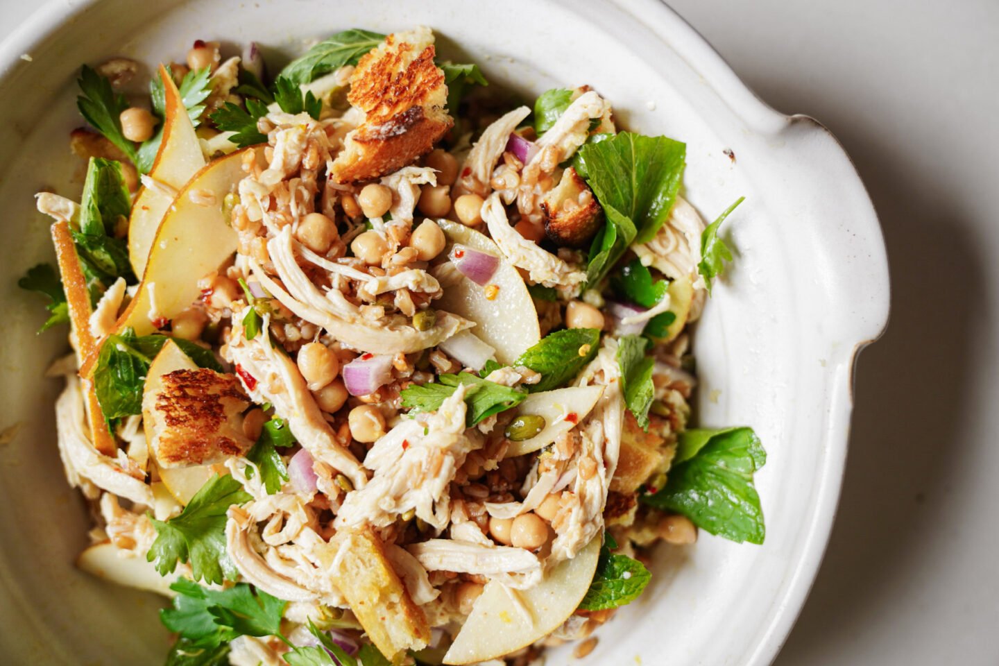 A bowl of salad with shredded chicken, chickpeas, sliced pears, red onions, fresh parsley, and toasted bread pieces, all mixed together on a white plate.