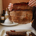 A person holds up a large, thin sheet of seasoned, cooked meat over a tray with more pieces in a kitchen setting; bottles and dishes are visible in the background.