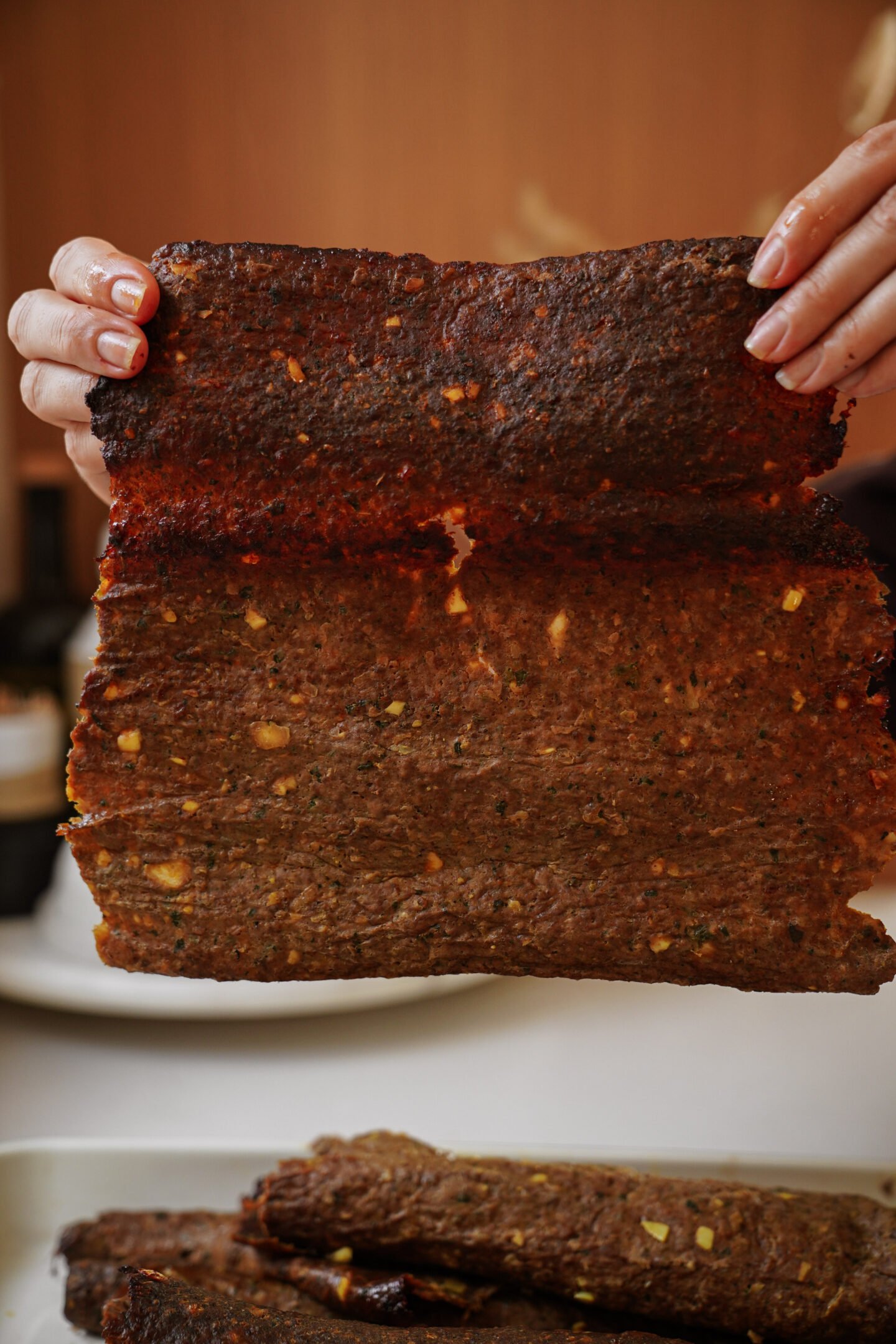A person holds up a large, thin sheet of seasoned, cooked meat, showing its texture and slightly uneven edges. The meat appears well-cooked with visible spices and small chunks of ingredients.