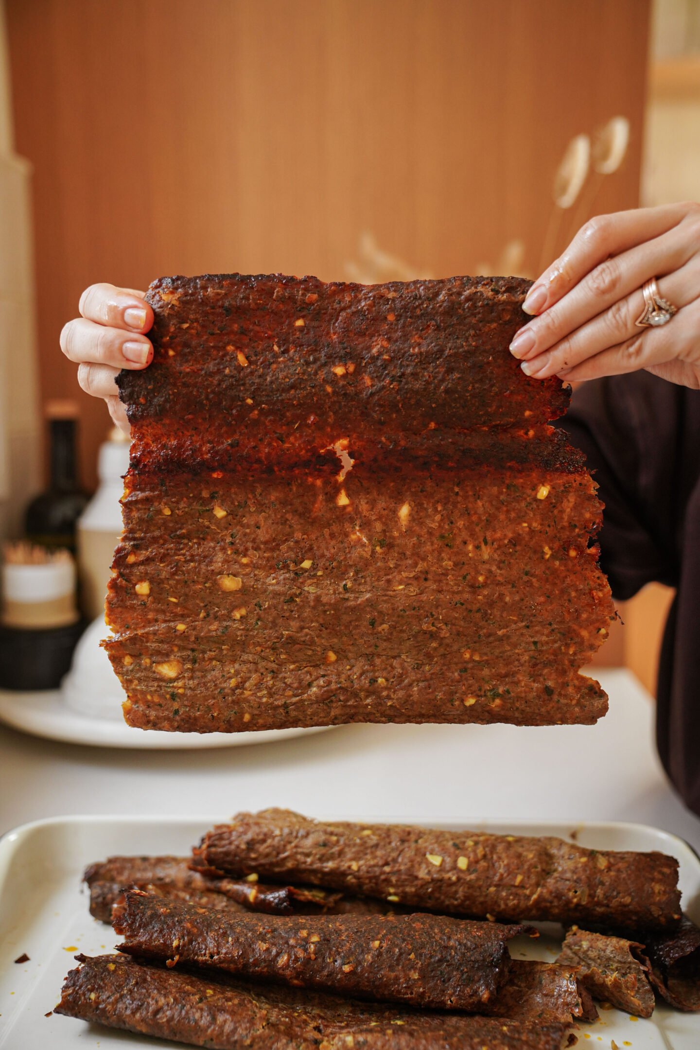 A person holds up a large, rectangular sheet of dark, crispy meat jerky over a tray with more pieces of jerky on it, in a kitchen setting.