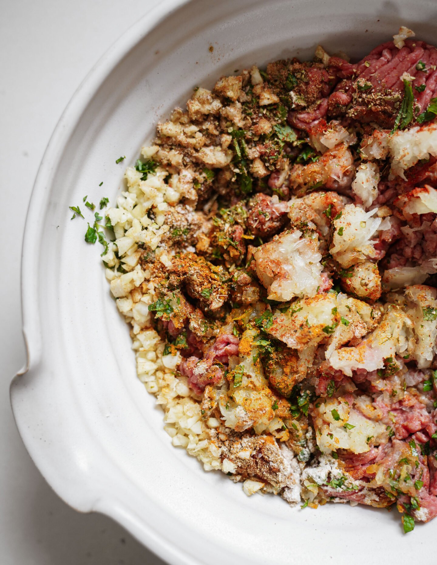 A close-up of a white bowl filled with raw ground meat, finely chopped onions, herbs, and spices, ready to be mixed for cooking.
