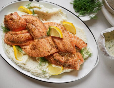 A braided baked salmon fillet garnished with dill and lemon slices, served on a bed of mashed potatoes, with extra dill and a bowl of creamy dill sauce on the side.
