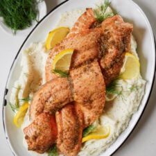 Plate of seasoned, baked salmon fillets topped with lemon slices and dill, served on a bed of creamy mashed potatoes, garnished with fresh dill sprigs. A small bowl of dill is nearby.