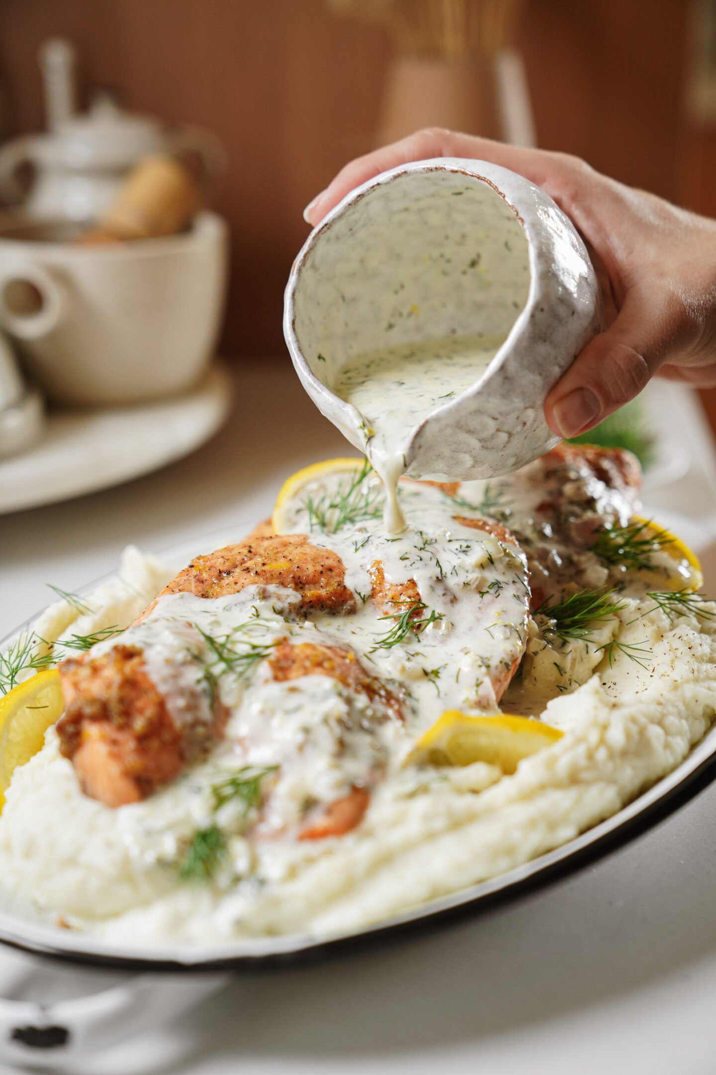 A hand pours creamy herb sauce over baked salmon fillets served on a bed of mashed potatoes, garnished with fresh dill and lemon slices. A teapot and cups are blurred in the background.