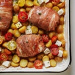 A baking tray with prosciutto-wrapped chicken, roasted baby potatoes, cherry tomatoes, and cubes of feta cheese, all seasoned with herbs and black pepper.