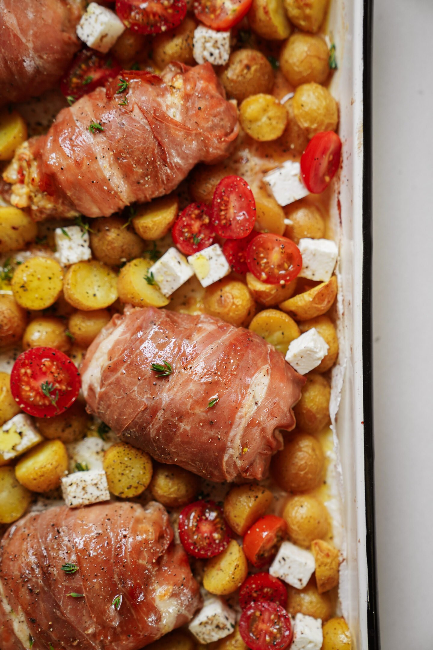 Baked prosciutto-wrapped chicken, golden baby potatoes, cherry tomato halves, and cubes of seasoned feta cheese on a white roasting tray.