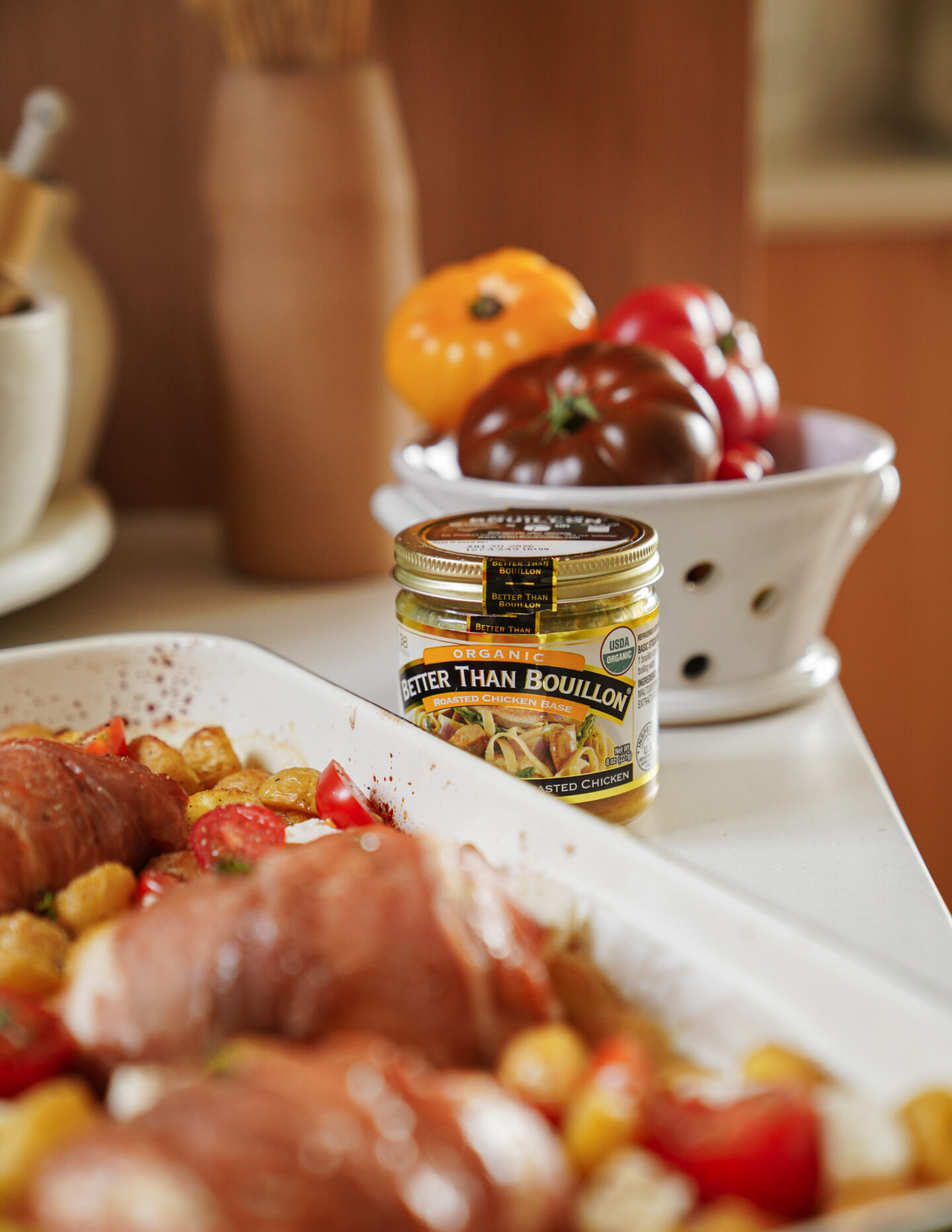 A jar of Better Than Bouillon roasted chicken base sits on a kitchen counter next to a bowl of heirloom tomatoes. In the foreground, a baking dish holds prepared meat and chopped vegetables.