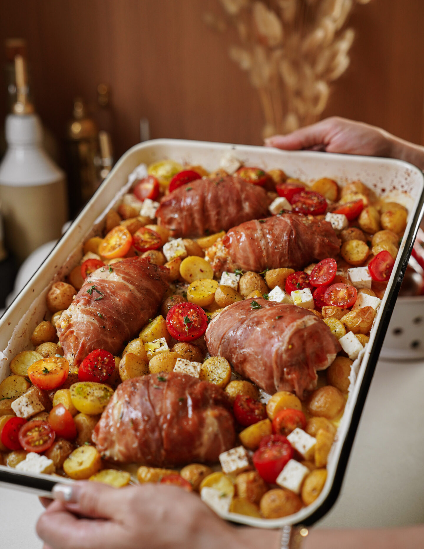 A baking tray filled with prosciutto-wrapped chicken, roasted baby potatoes, cherry tomatoes, and cubes of feta cheese, held by hands in a kitchen setting.
