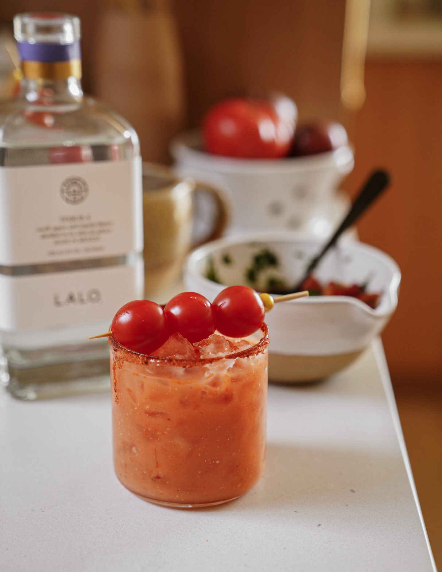 A glass of tomato-based cocktail with ice and a chili rim, garnished with three cherry tomatoes on a skewer, sits on a counter near a bottle of tequila and bowls of chopped herbs and tomatoes.