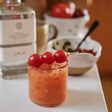 A cocktail with a salted rim and ice sits on a counter, garnished with three cherry tomatoes on a skewer. Behind it are a bottle of LALO tequila, a bowl of tomatoes, and a bowl with herbs and a spoon.