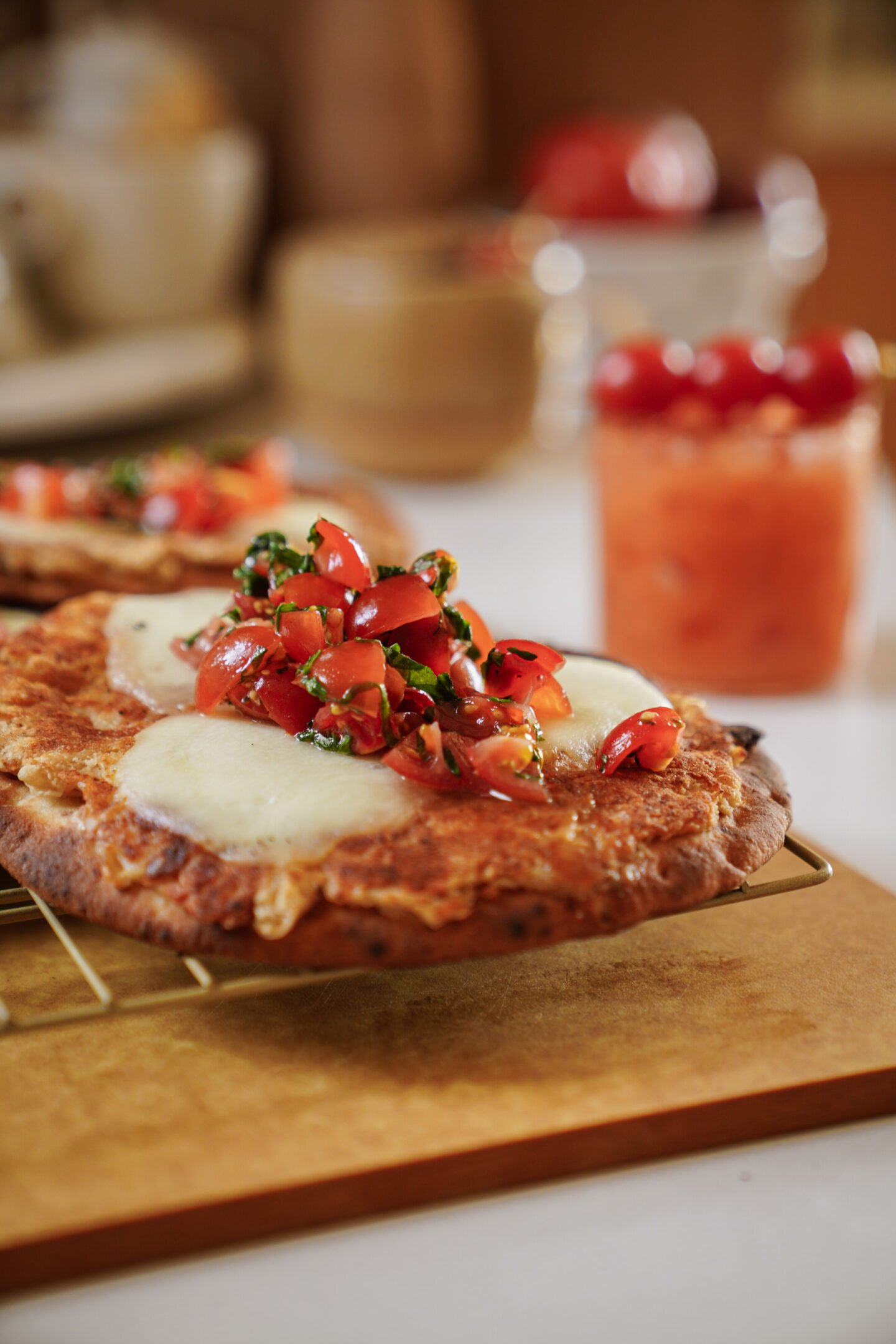A close-up of a flatbread topped with melted cheese and a fresh tomato-basil mixture, resting on a cooling rack. Other blurred food items and a glass of orange beverage are visible in the background.