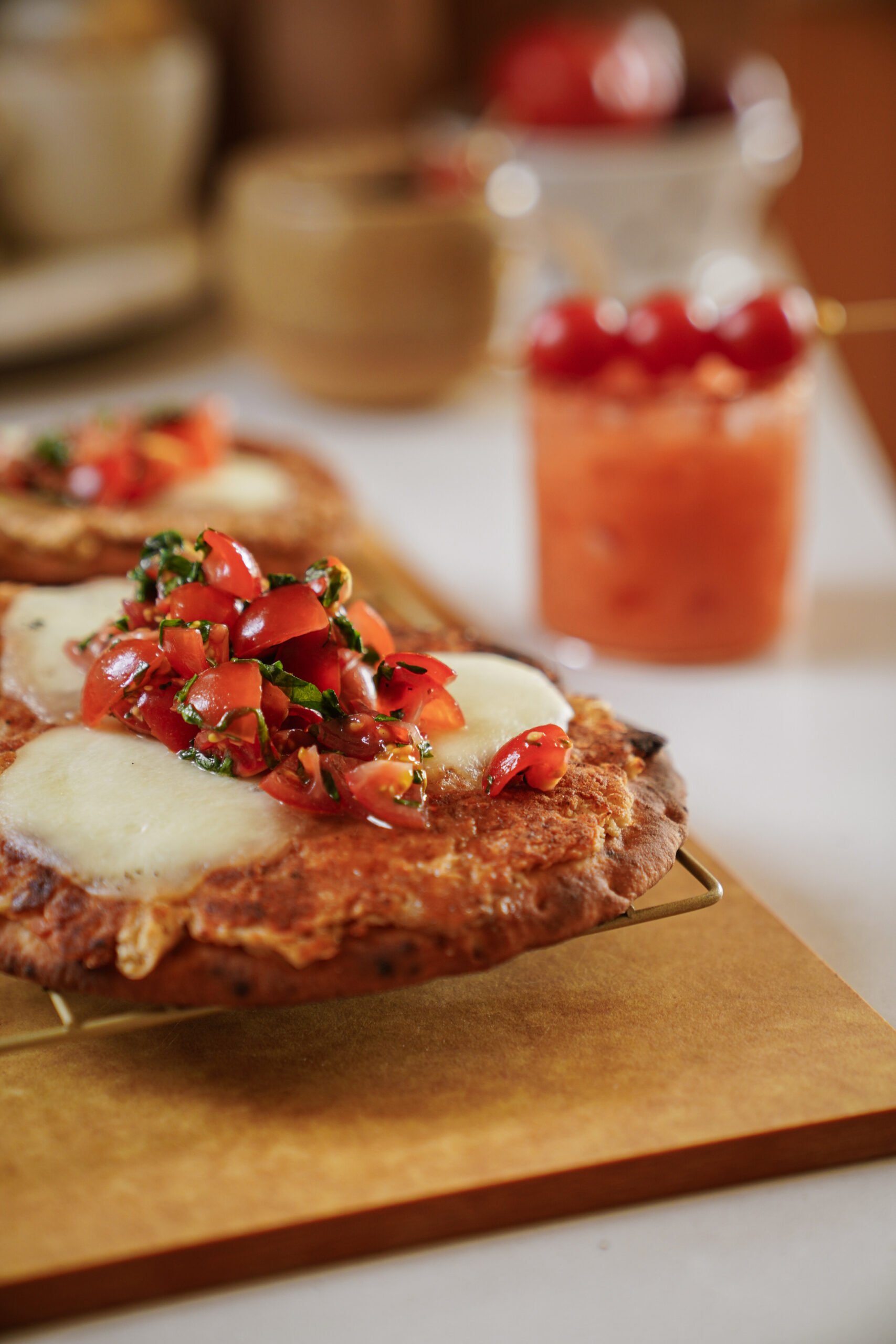 A close-up of flatbread pizzas topped with melted mozzarella and fresh tomato-basil bruschetta, set on a cooling rack. In the background, a glass with a pink drink and skewered cherries is visible.