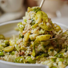 A close-up of a spoonful of grain salad with Brussels sprouts, barley, and diced vegetables being lifted from a white bowl. The dish appears fresh and healthy.