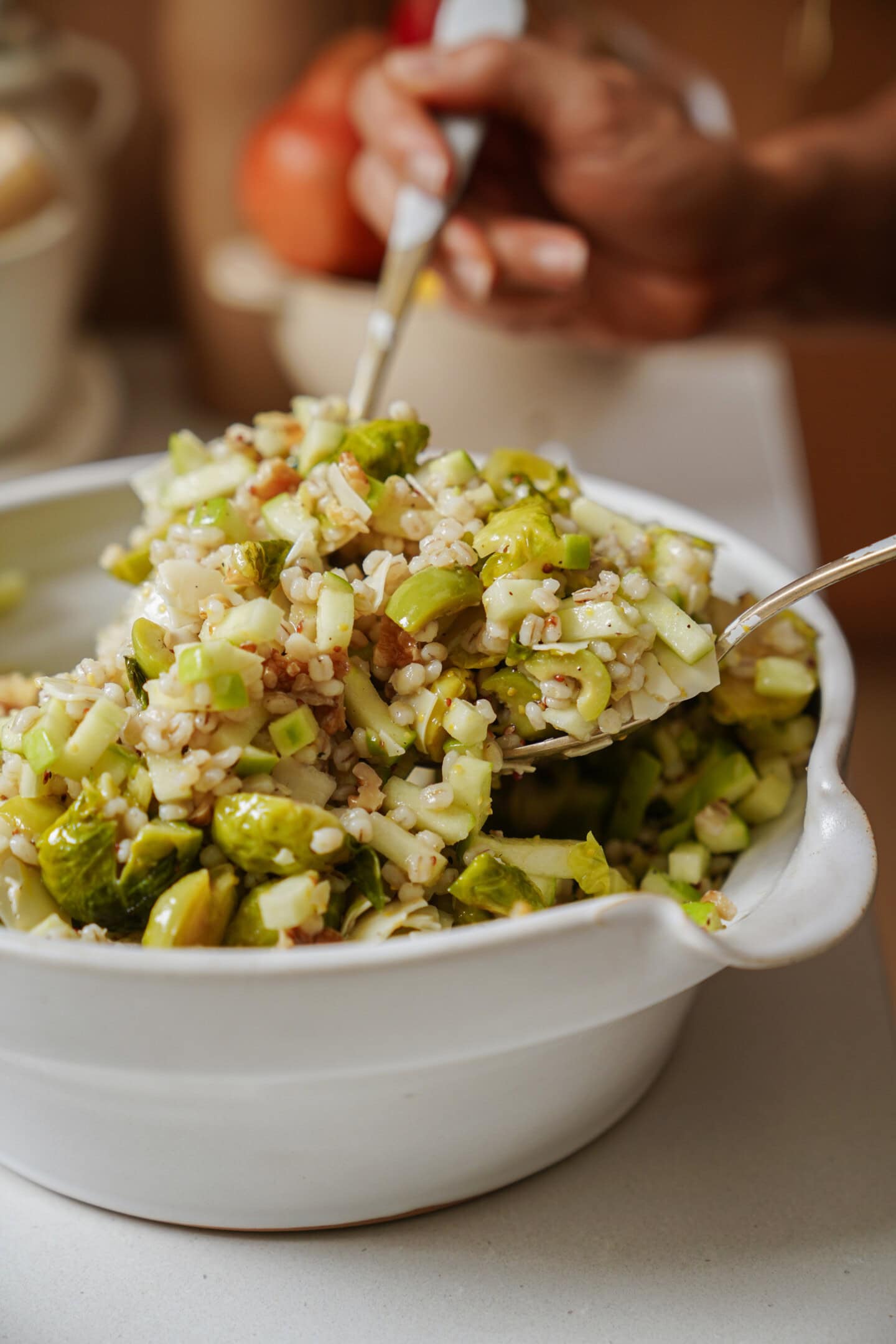 A close-up of a hand serving a barley salad with Brussels sprouts, chopped celery, and possibly nuts from a white bowl using a large spoon and fork.