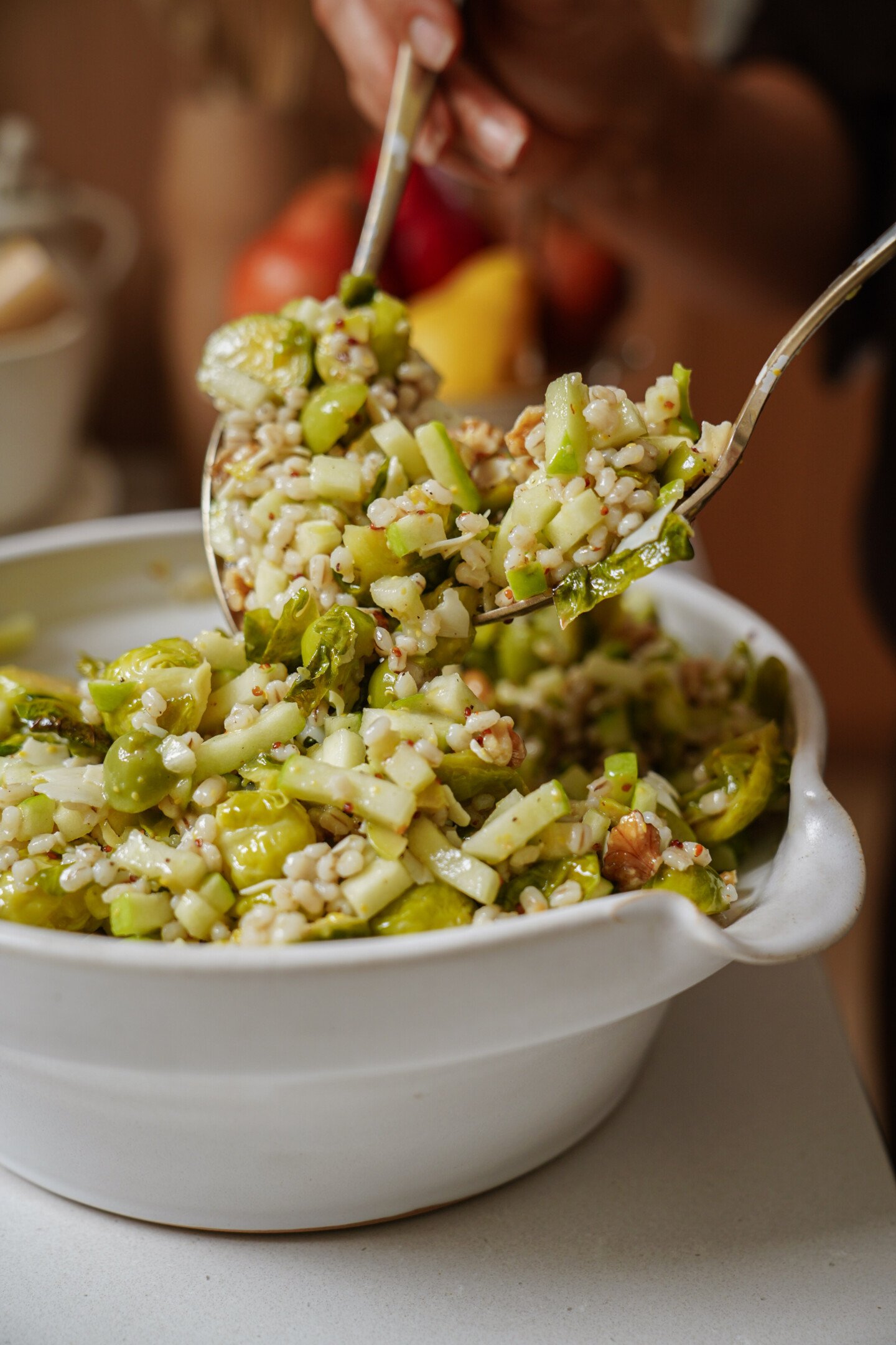 A person scoops a serving of grain and vegetable salad from a white bowl using two serving utensils. The salad contains barley, sliced vegetables, and herbs.