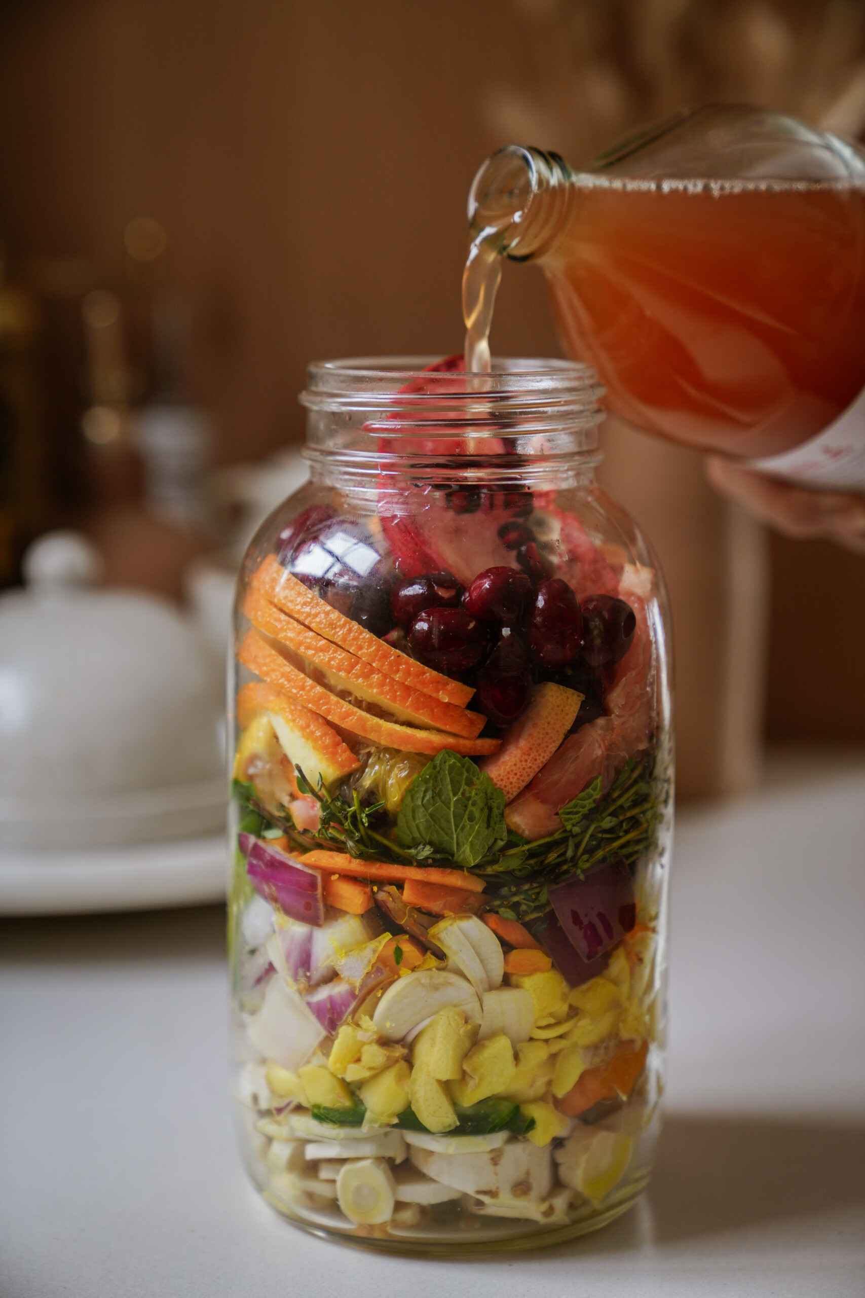 A hand pours liquid from a glass bottle into a large mason jar filled with layers of colorful fruits, vegetables, and herbs on a kitchen counter.