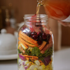 A glass jar filled with colorful chopped fruits, vegetables, and herbs is being topped with a light orange liquid poured from a bottle. The ingredients are layered in a visually appealing way.