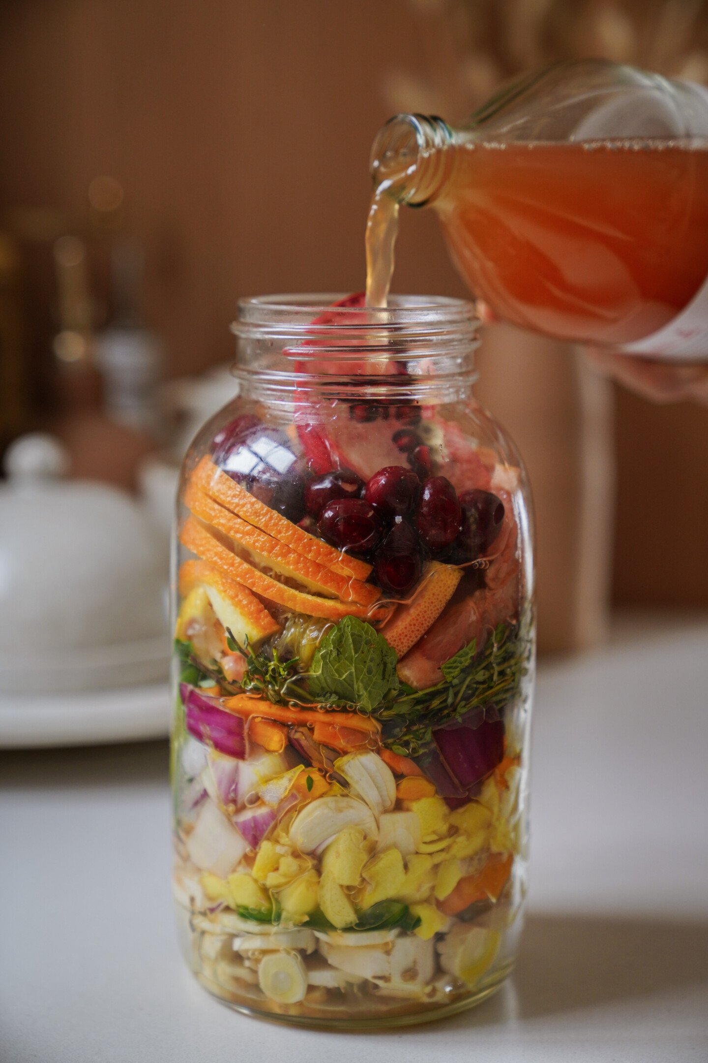 A hand pours pinkish liquid from a glass bottle into a large mason jar filled with colorful layers of chopped fruits, vegetables, and herbs on a kitchen counter.