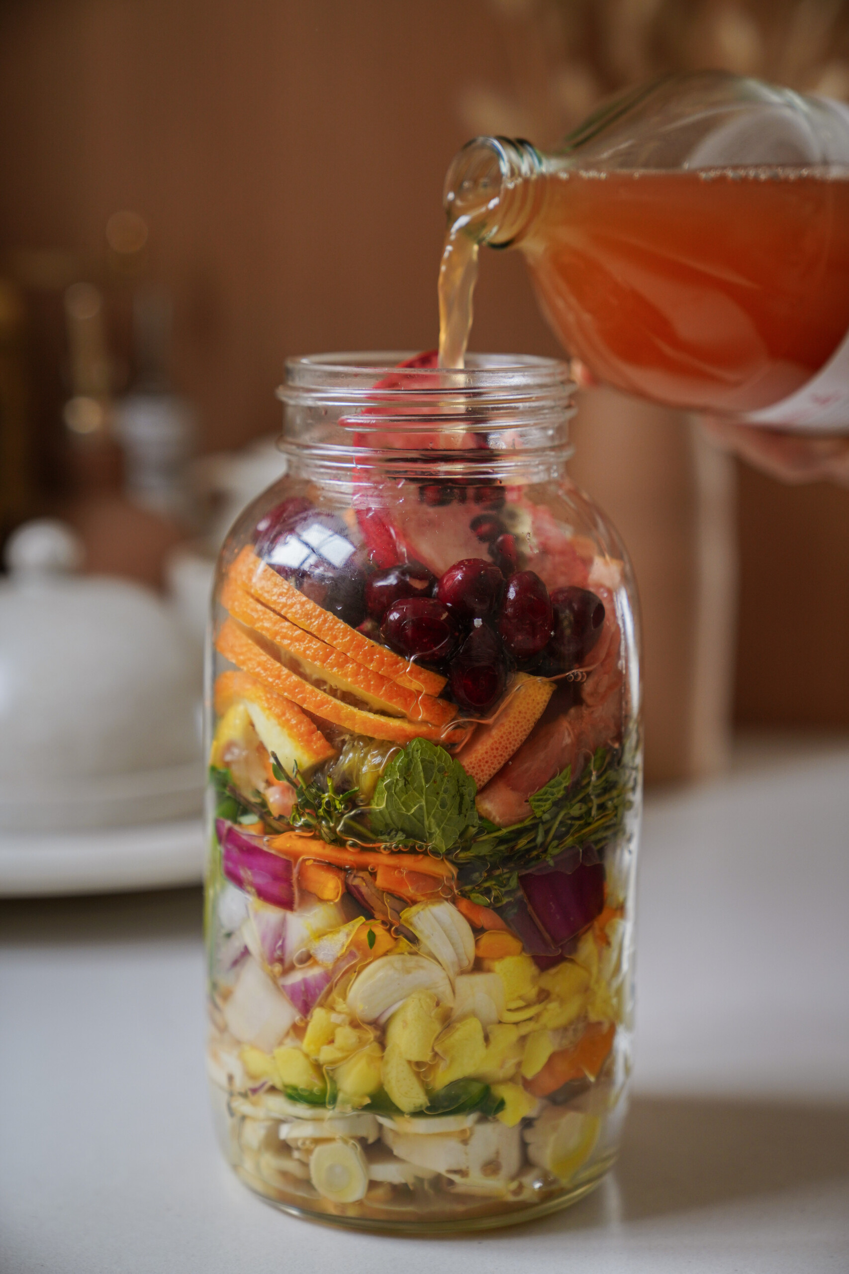 A hand pours pinkish liquid from a glass bottle into a large mason jar filled with colorful layers of chopped fruits, vegetables, and herbs on a kitchen counter.