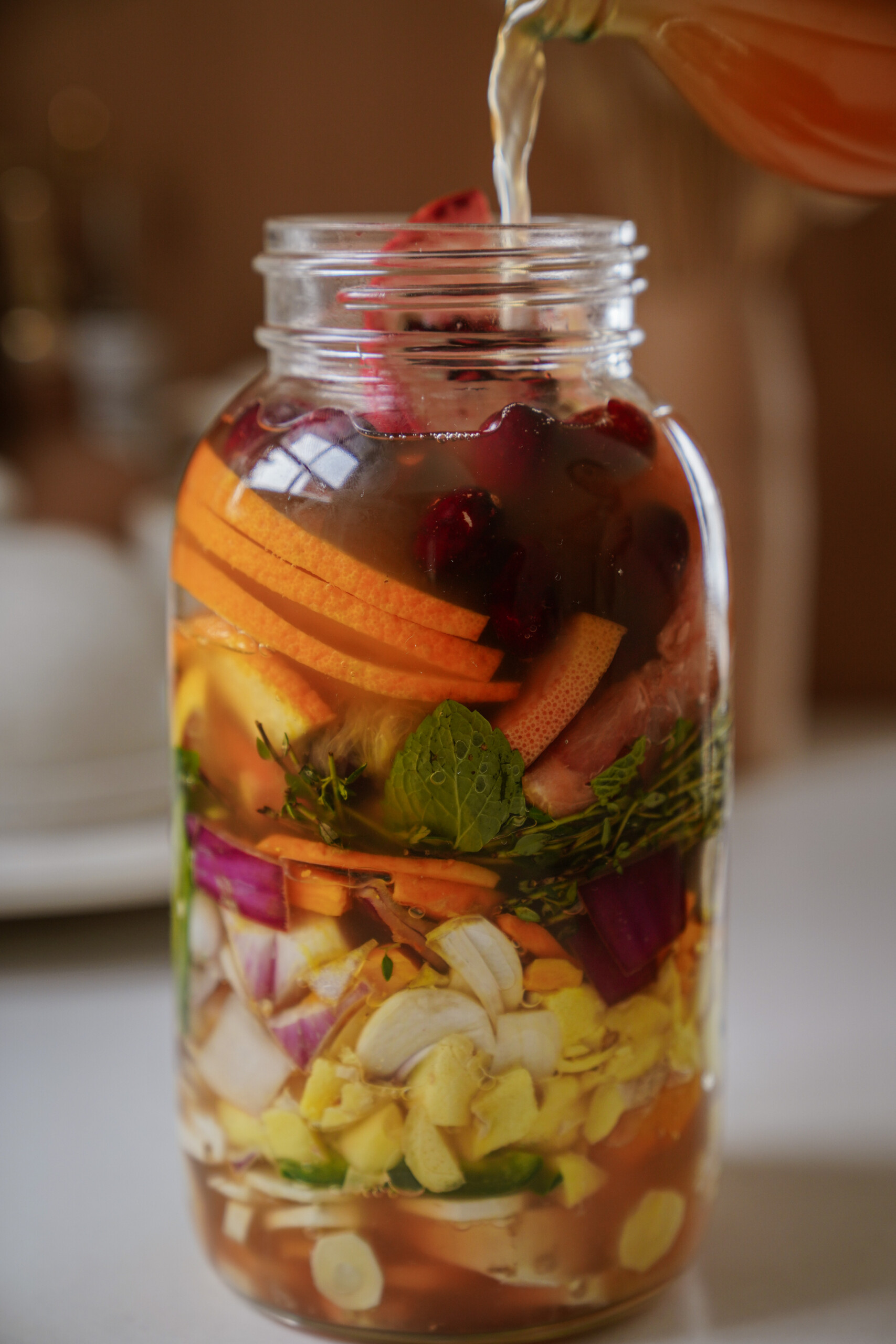 A glass jar filled with colorful sliced vegetables, herbs, and spices, as liquid is being poured over them from a bottle, creating a vibrant layered mixture on a kitchen counter.
