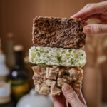 A hand holds three stacked rectangular food bars: a brown crunchy bar on top, a white rice and green herb bar in the middle, and a chunky beige bar with visible pieces on the bottom.