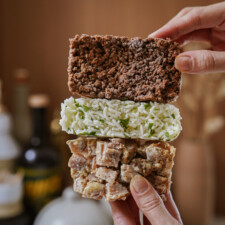 A hand holds three stacked rectangular food bars: a brown crunchy bar on top, a white rice and green herb bar in the middle, and a chunky beige bar with visible pieces on the bottom.