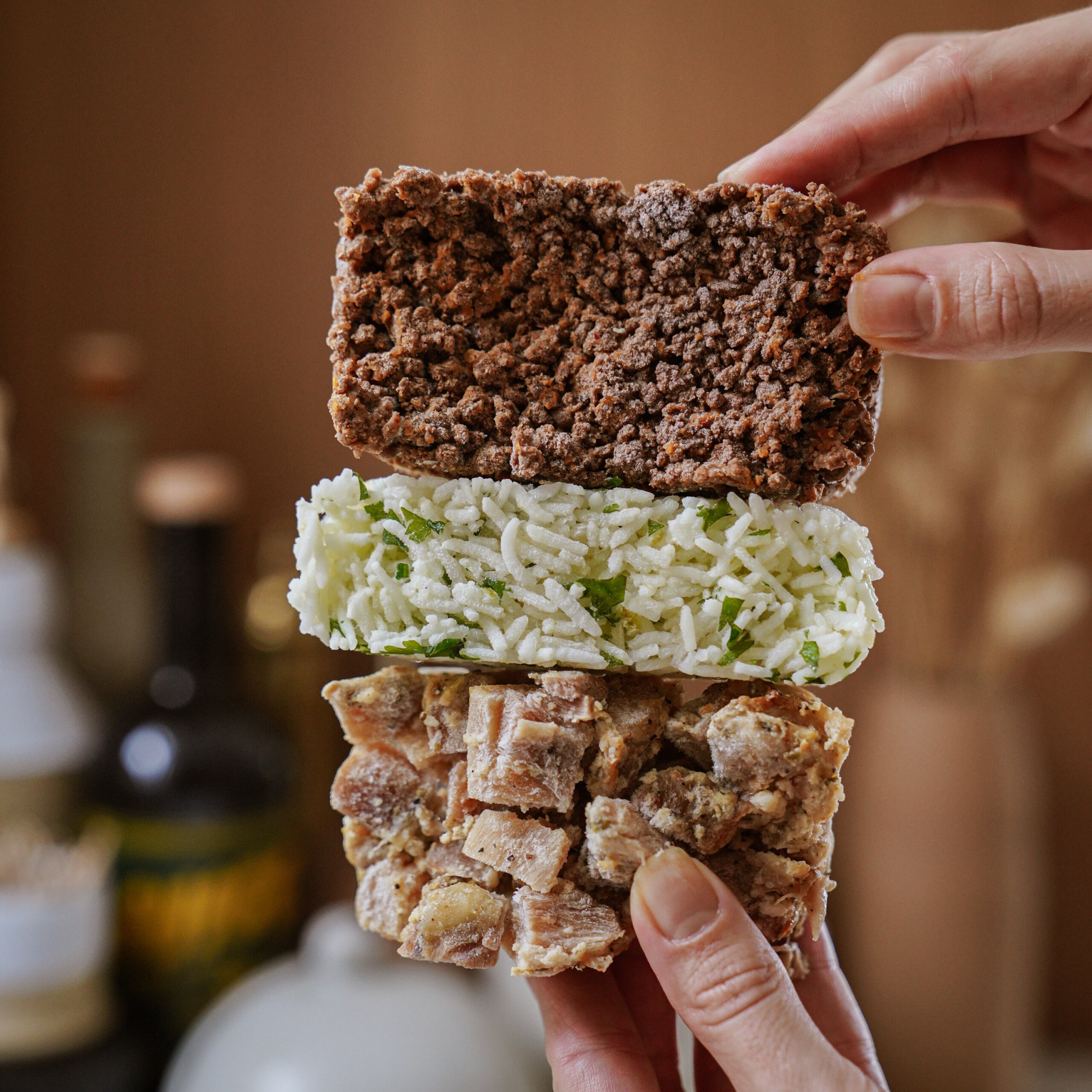A hand holds three stacked rectangular food bars: a brown crunchy bar on top, a white rice and green herb bar in the middle, and a chunky beige bar with visible pieces on the bottom.