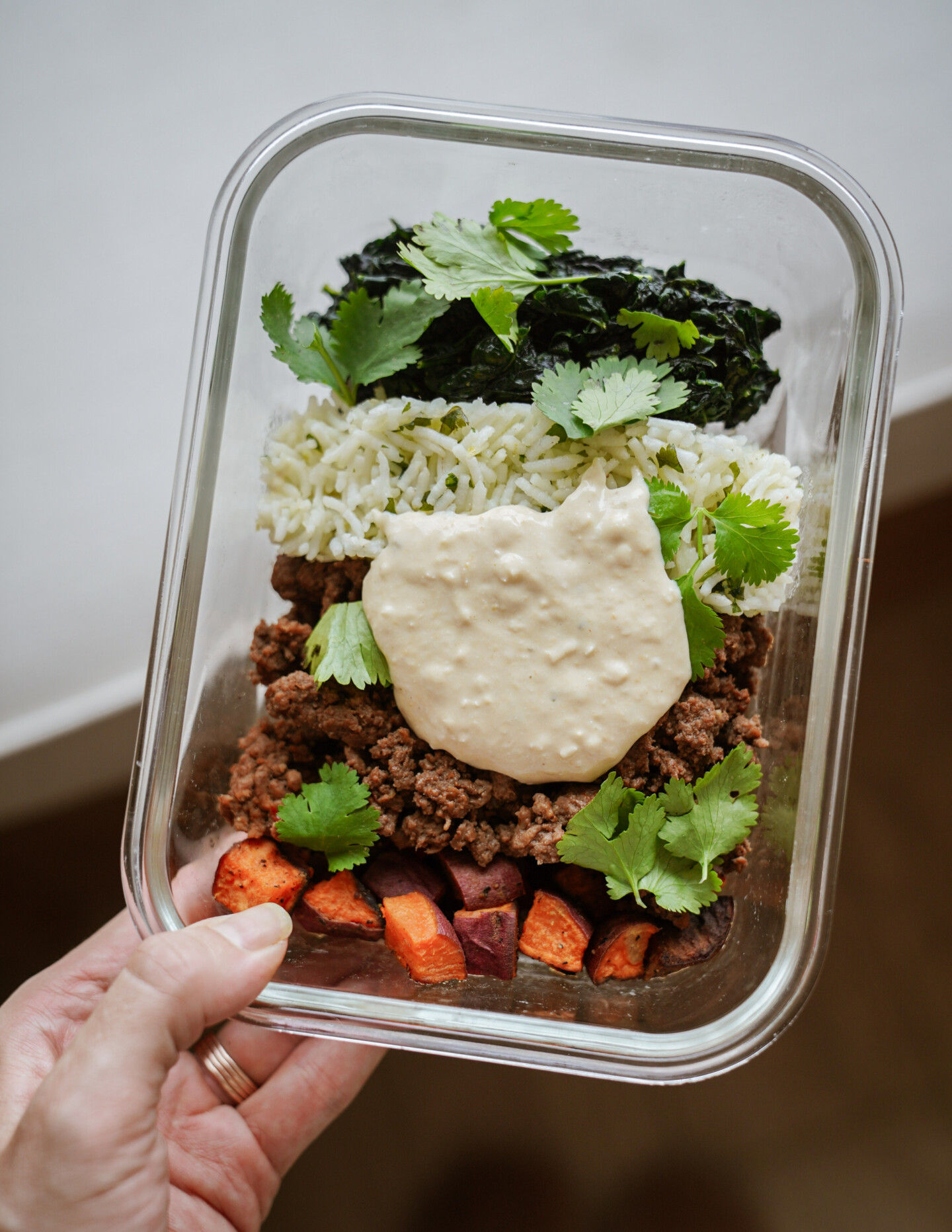 A hand holds a glass container filled with cooked ground beef, rice, roasted sweet potatoes, greens, a dollop of creamy sauce, and garnished with fresh cilantro leaves.