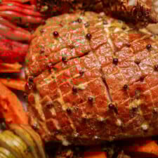 Close-up of a glazed, baked ham studded with whole cloves, scored in a diamond pattern, surrounded by colorful vegetables and garnishes.