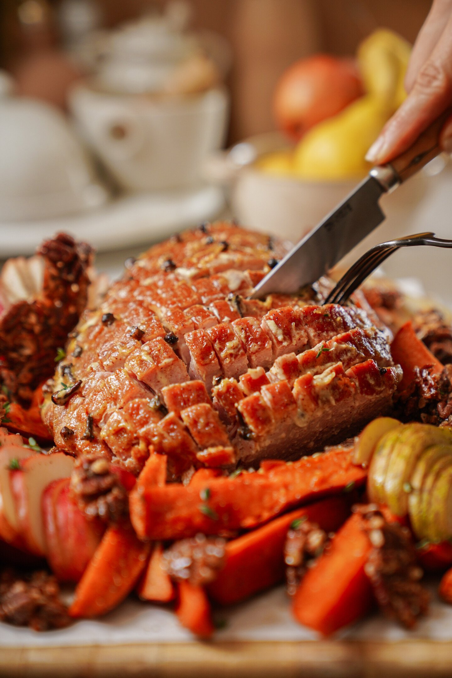 A hand slices a roasted ham with a knife and fork. The ham is glazed and surrounded by roasted vegetables, including carrots, apples, and walnuts. The background shows blurred dishes and fruit.