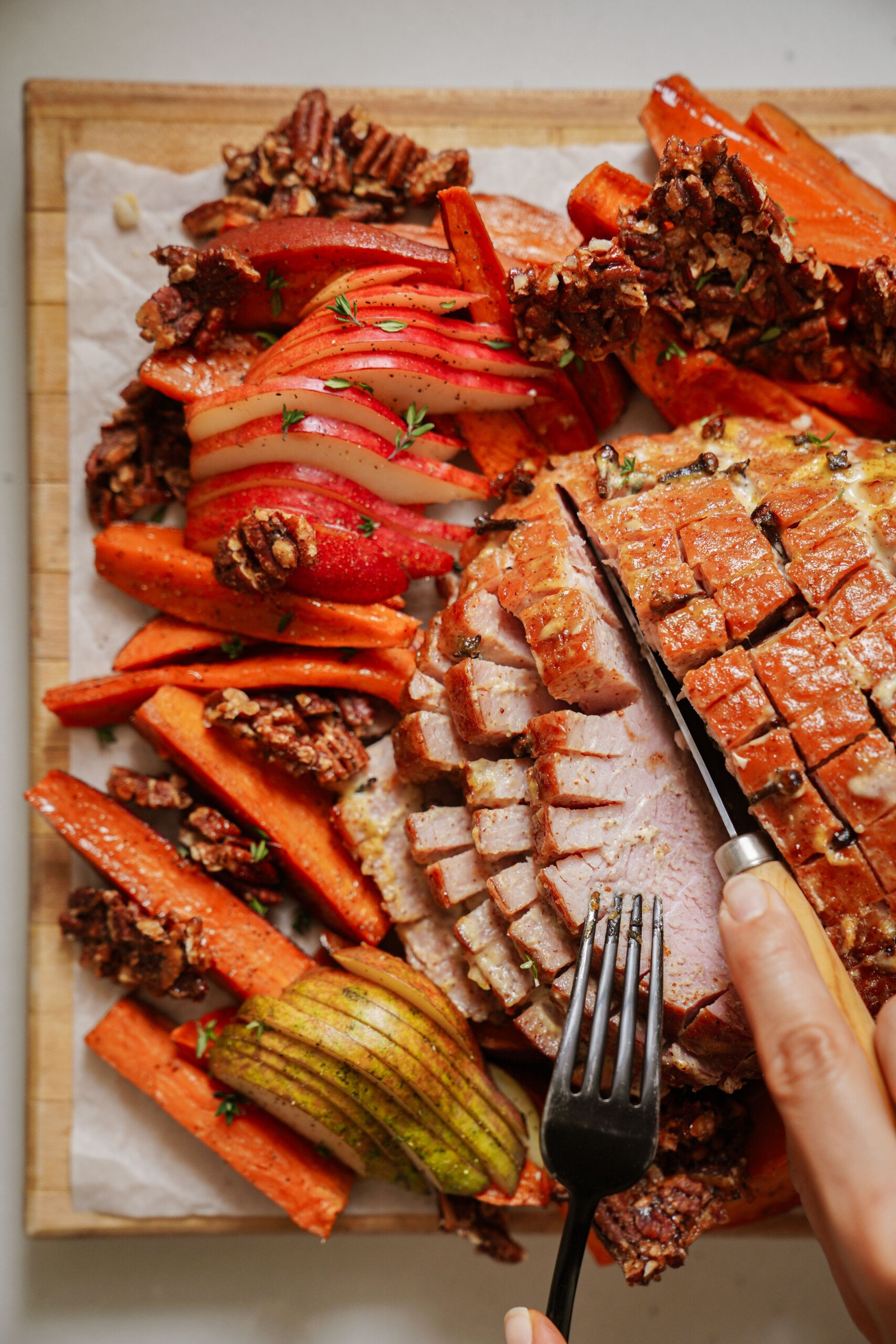 A hand slices glazed ham on a wooden board, surrounded by roasted carrots, sliced pears, sliced red apples, and clusters of candied pecans.