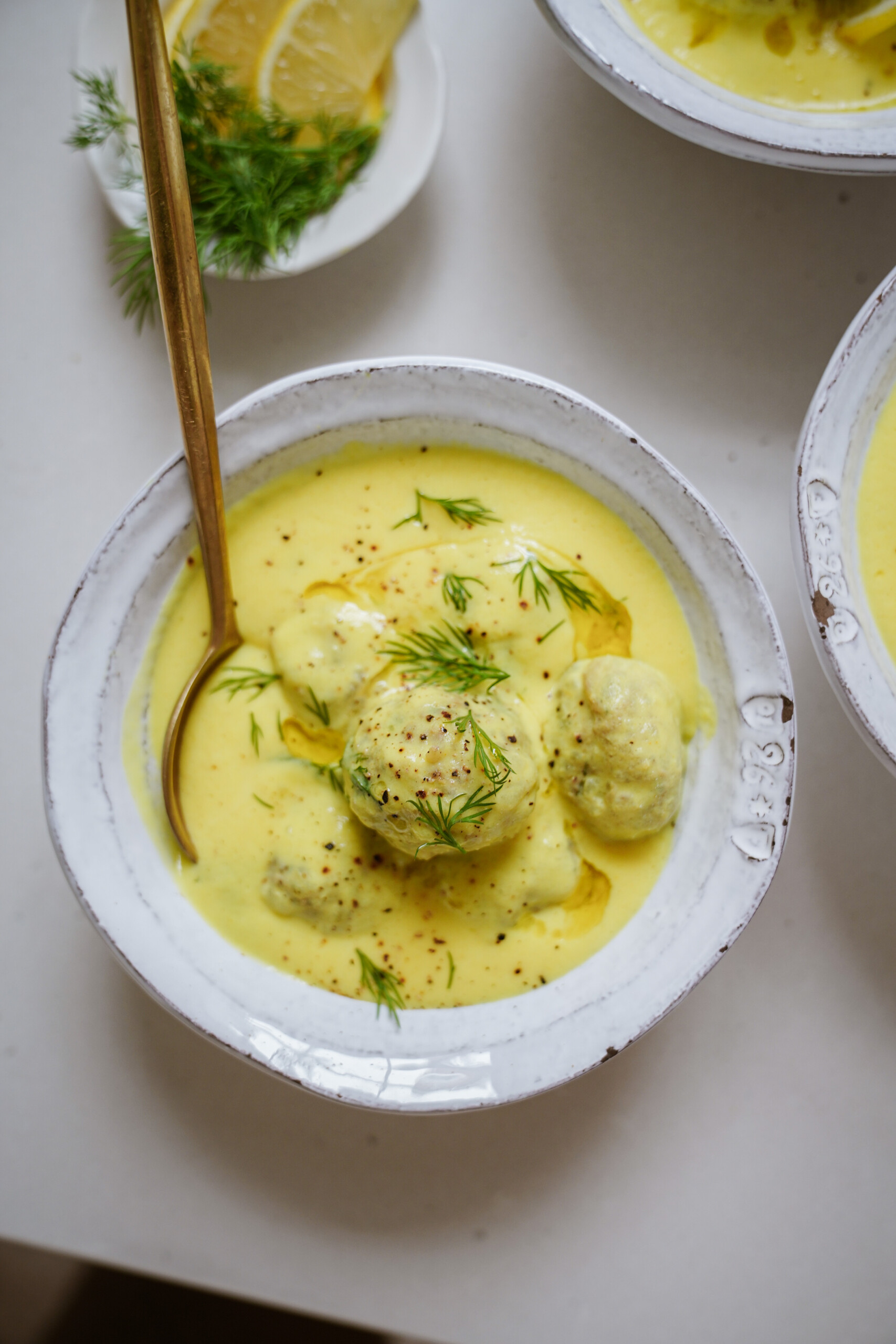 A bowl of creamy yellow soup with meatballs, garnished with fresh dill and black pepper, sits on a white surface with a gold spoon. Lemon slices and dill are visible on a small plate in the background.