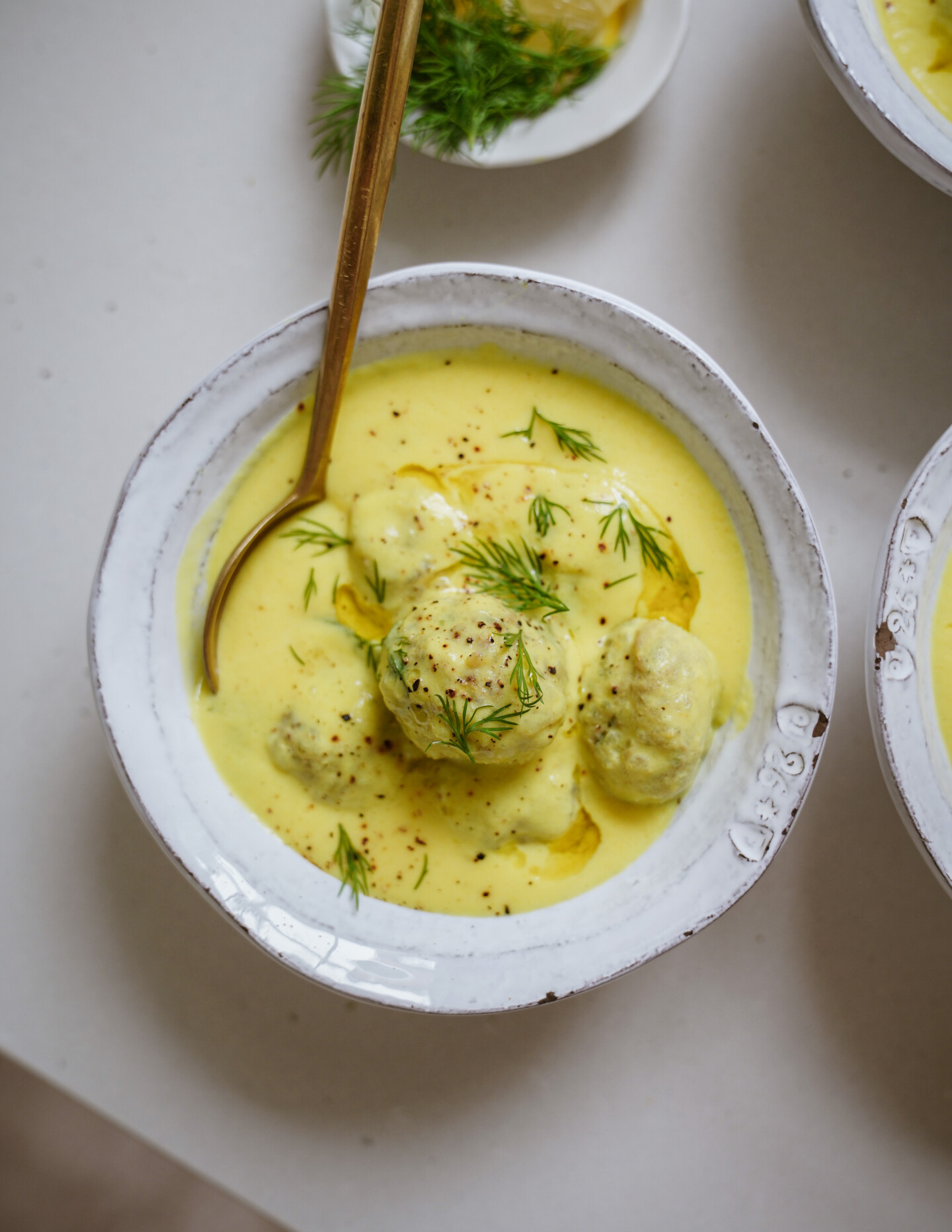 A bowl of creamy yellow soup with dumplings, garnished with fresh dill and black pepper, with a spoon resting inside. The bowl sits on a light-colored surface.