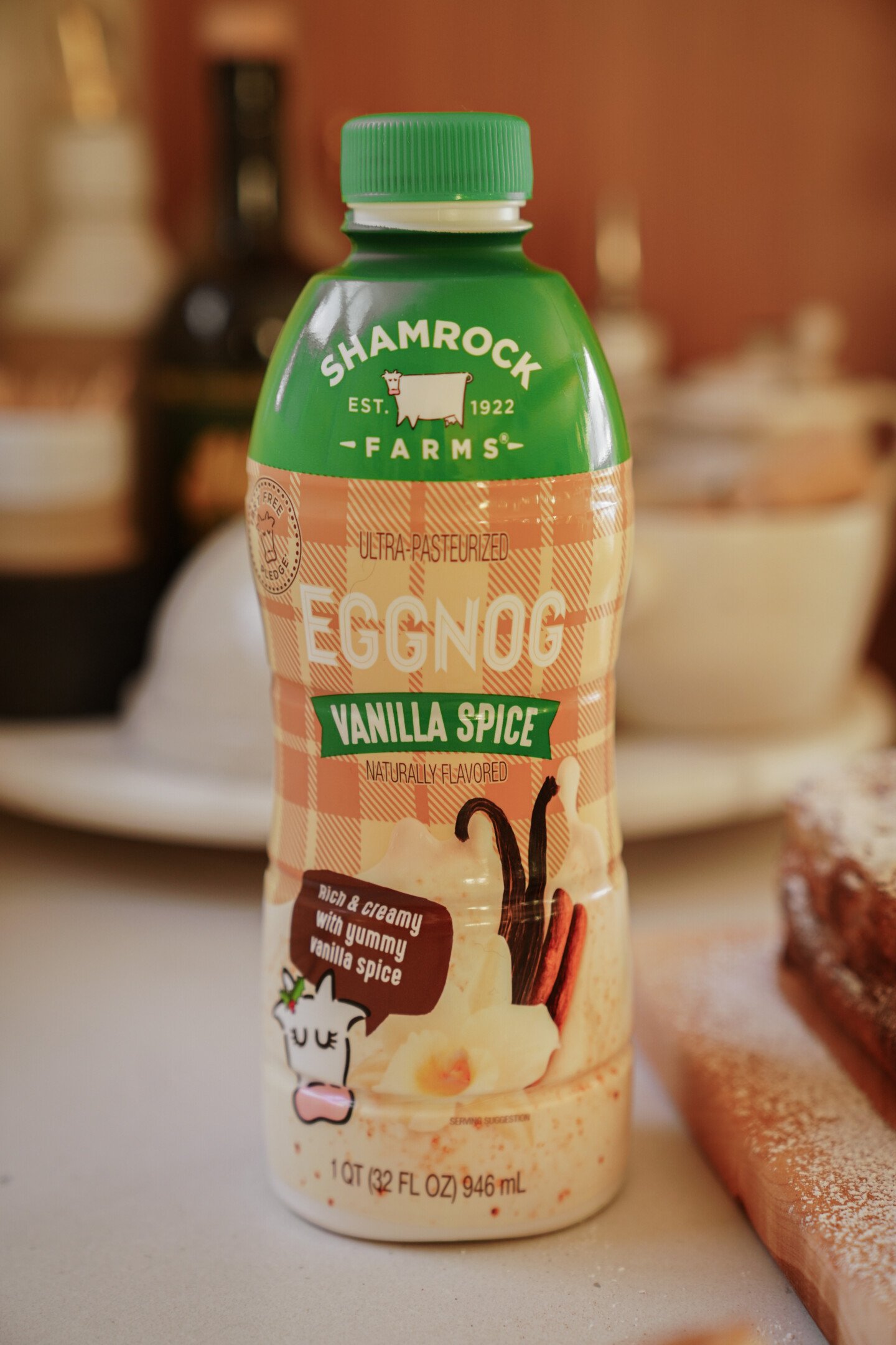 A 1-quart bottle of Shamrock Farms Vanilla Spice Eggnog with a green cap sits on a counter. The label features images of vanilla beans, a vanilla flower, and a cartoon cow.