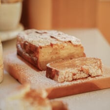 A sliced loaf of apple cake dusted with powdered sugar sits on a wooden cutting board, with a soft, golden-brown crust and visible apple layers inside.