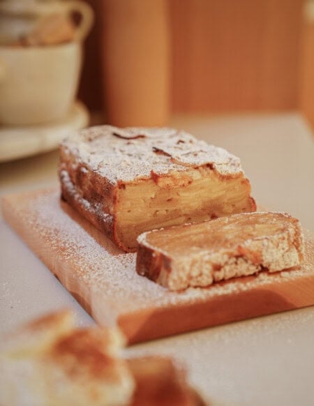 A rectangular apple cake dusted with powdered sugar sits on a wooden cutting board. One slice is cut and laid in front, revealing layers of apple inside the cake. The background is softly blurred.