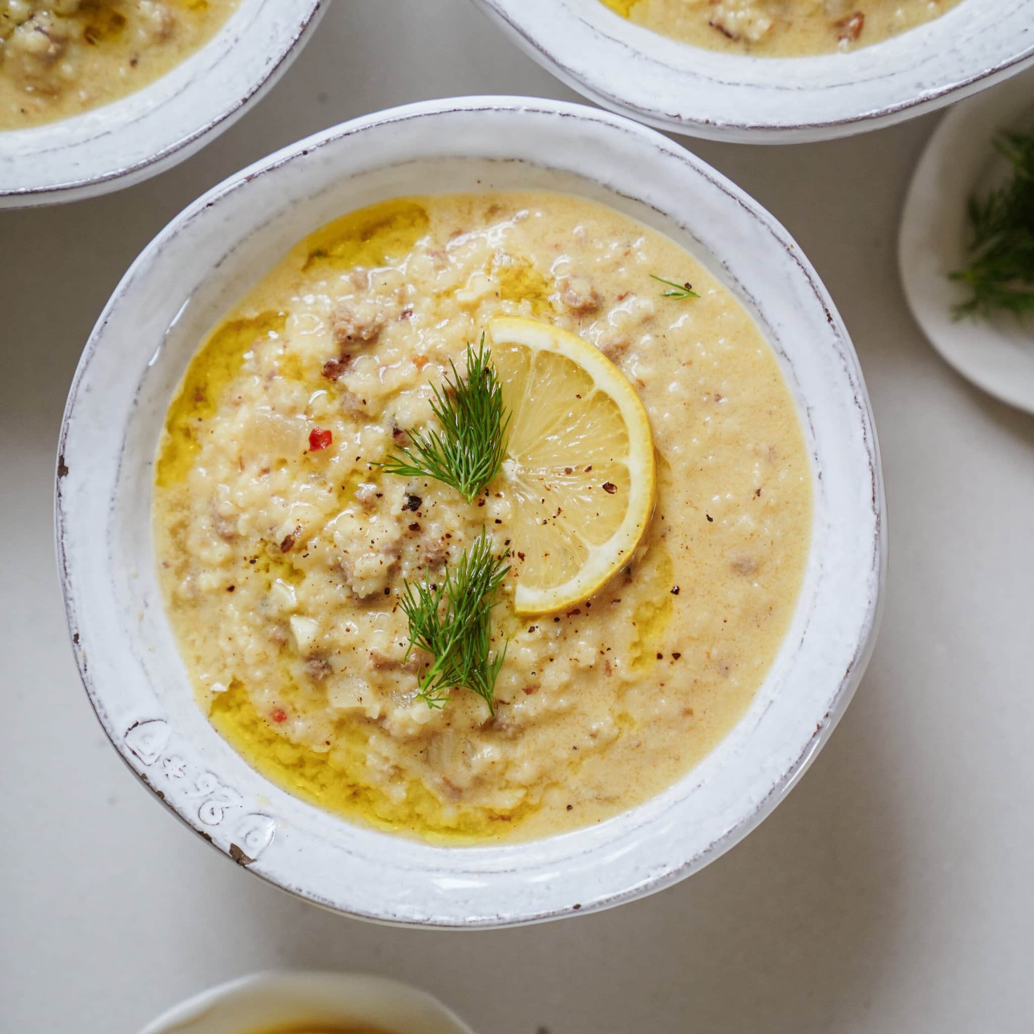 A bowl of creamy soup topped with a lemon slice and fresh dill, with a drizzle of oil on the surface. Two additional bowls and a small plate with dill are partially visible in the background.