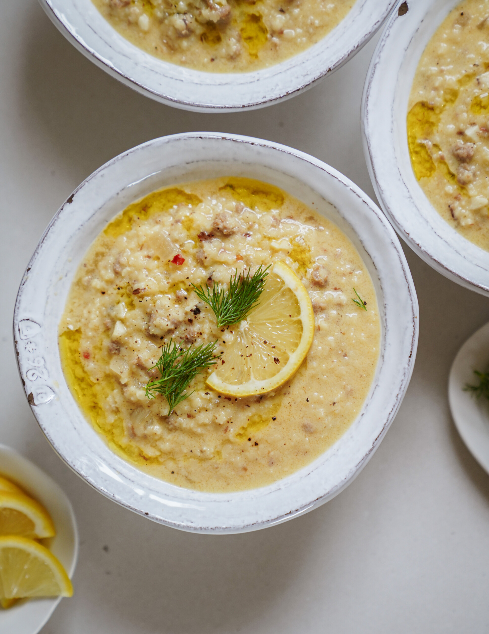 A bowl of creamy soup topped with a lemon slice and fresh dill, served in a white ceramic bowl, with other similar bowls and lemon wedges nearby.