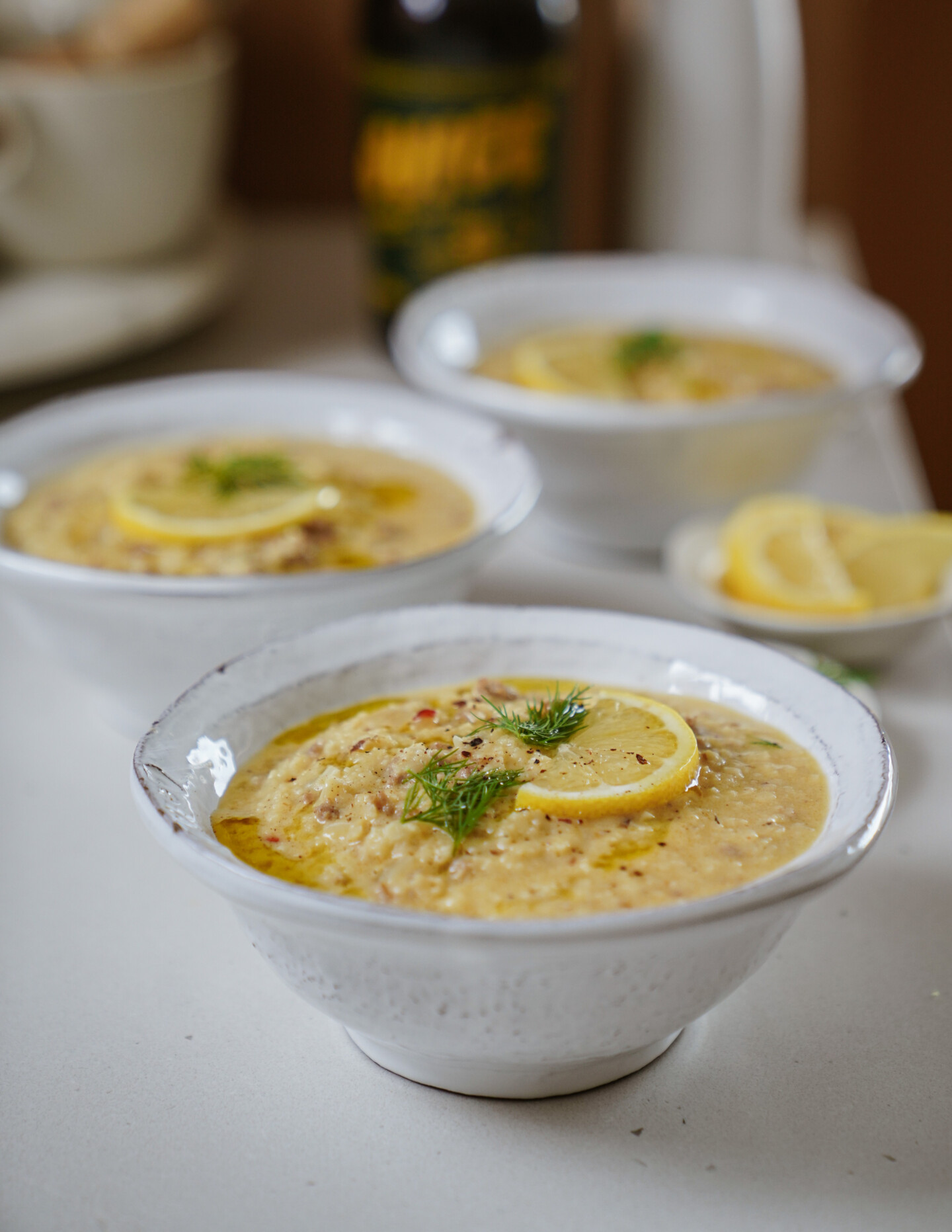 Three white bowls of creamy soup are garnished with lemon slices and fresh dill, sitting on a light surface. A bottle and a small dish of lemon wedges are blurred in the background.