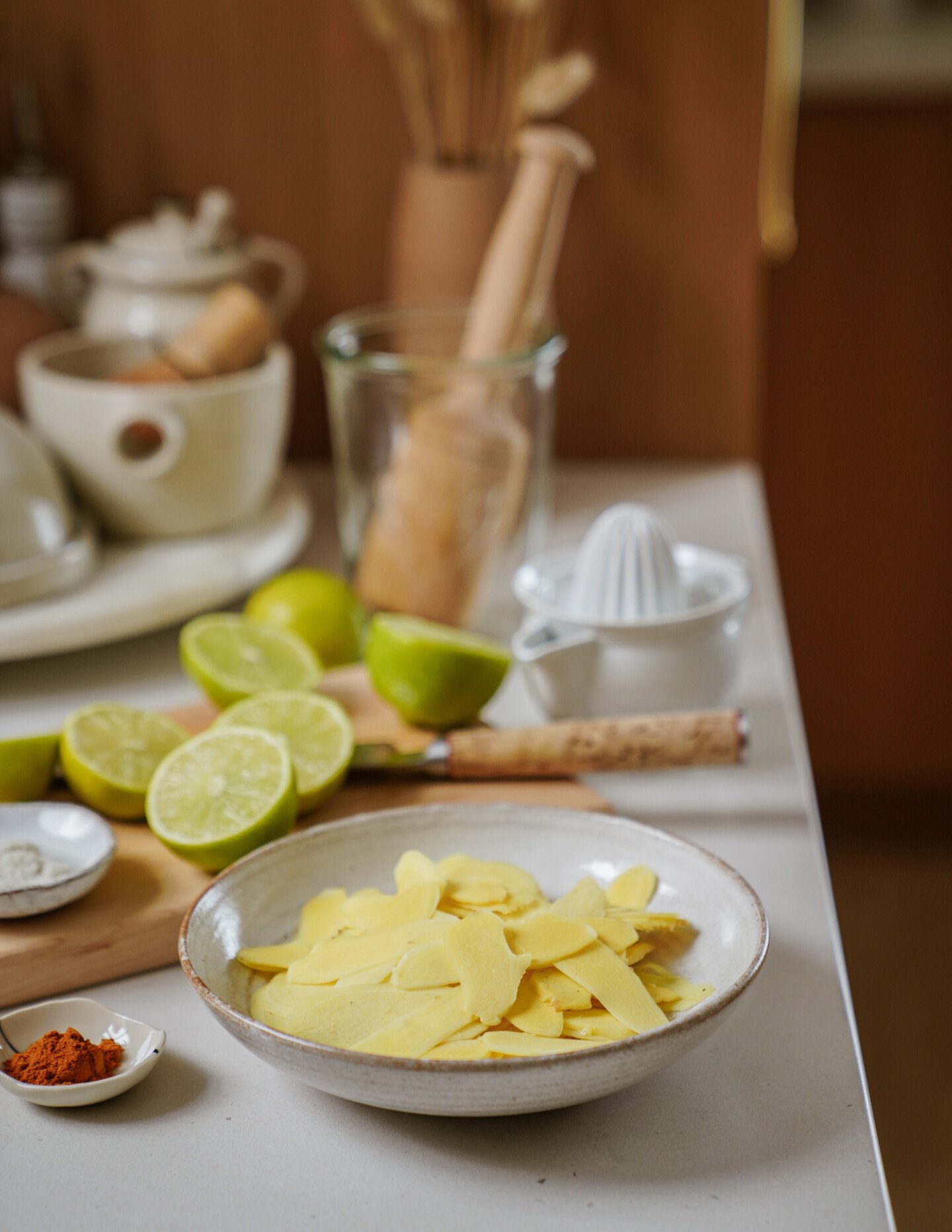 A bowl of thinly sliced ginger sits on a kitchen counter beside cut limes, a wooden citrus juicer, a glass jar, a teapot, and small dishes of salt and red spice. The background shows more kitchen items.